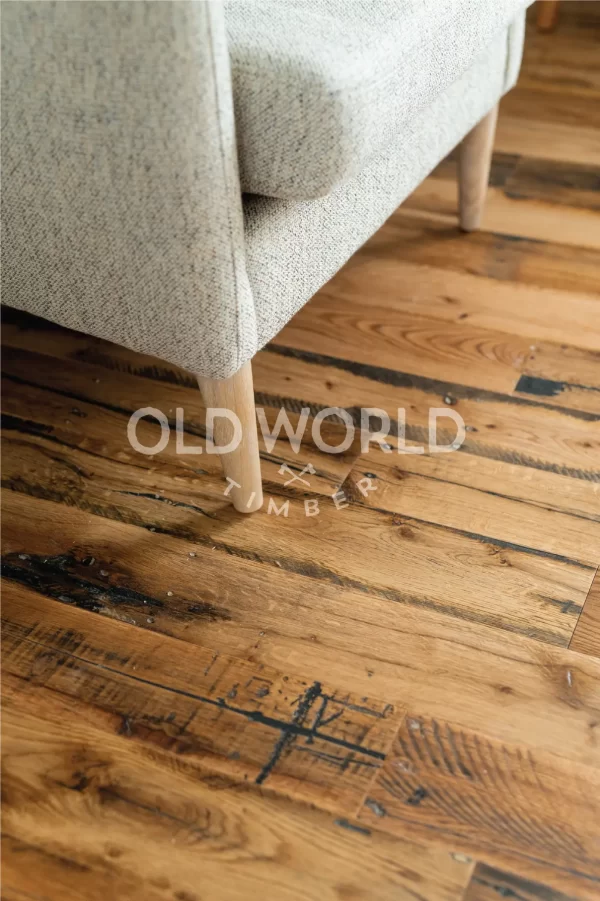 Close-up of reclaimed industrial oak floor beneath a beige fabric armchair, displaying dark character streaks and wear patterns from historic use.