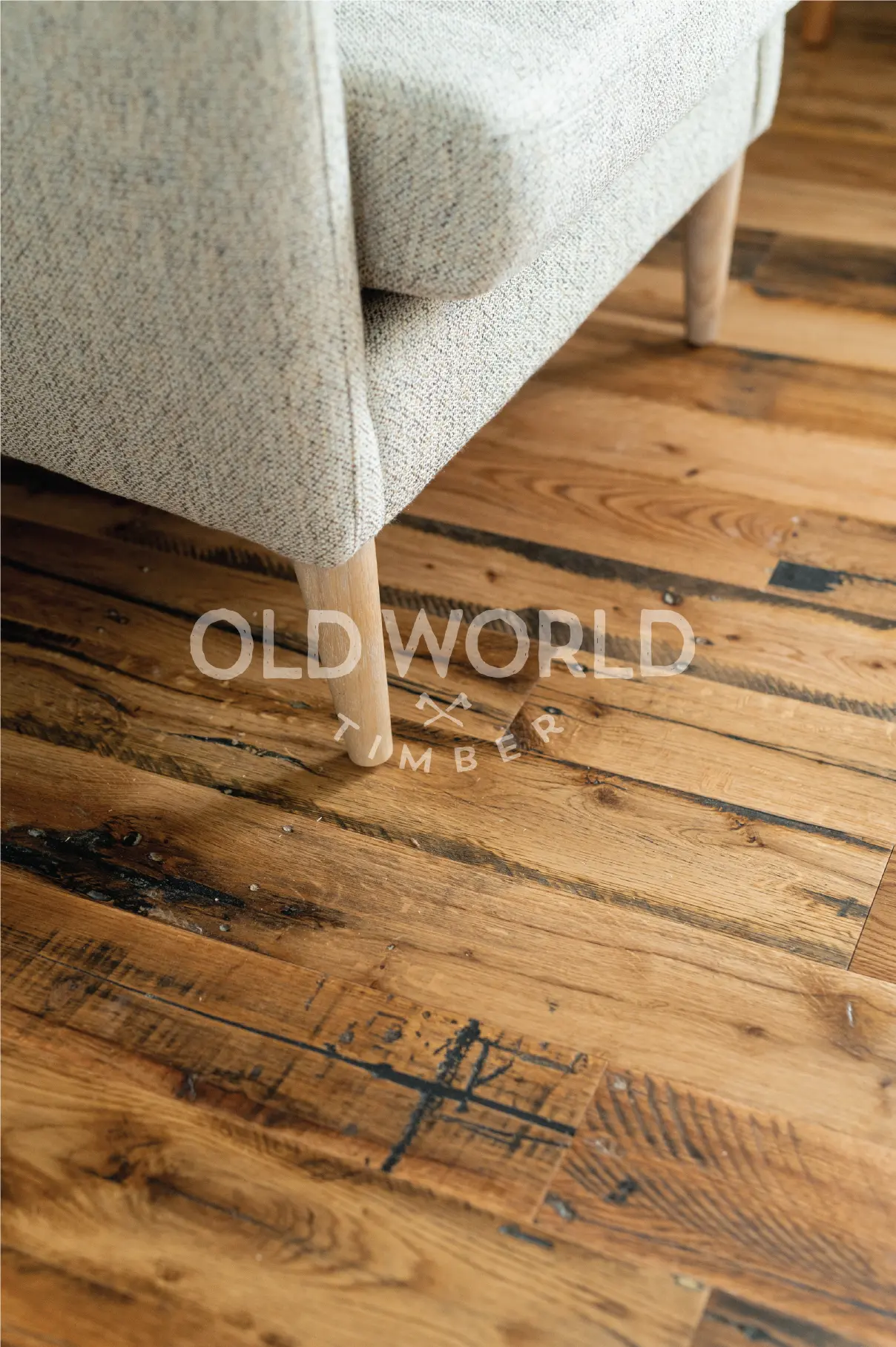 Close-up of reclaimed industrial oak floor beneath a beige fabric armchair, displaying dark character streaks and wear patterns from historic use.